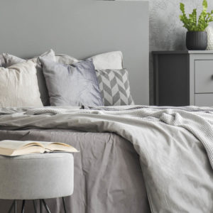 Open book placed on pouf standing next to double bed with grey b