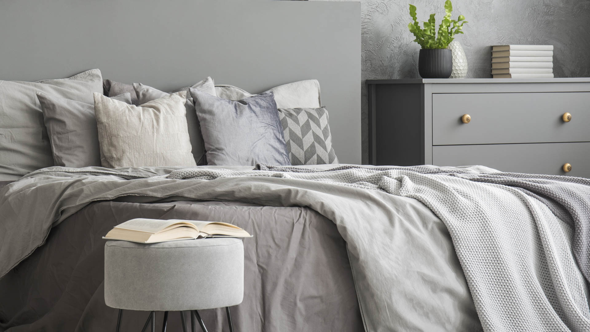 Open book placed on pouf standing next to double bed with grey b